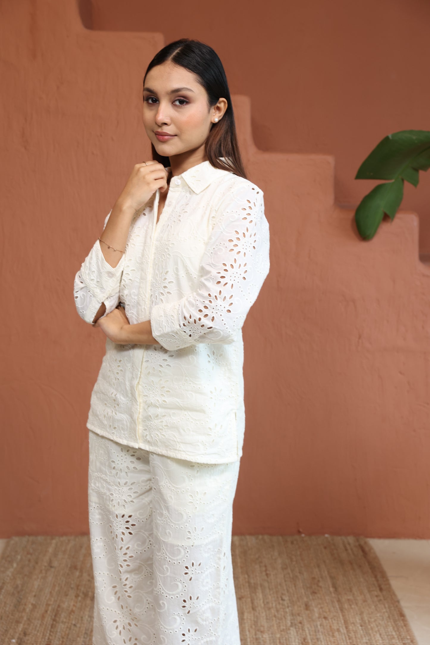 Woman in a white outfit sitting on a chair against a brown wall. White Top With Trouser Schiffli Co-Ords, Printed Co-Ords Set, Printed Palazzos Set, printed co-ords, Floral printed, co-ord set, Shirt and palazzo set, women’s co-ords, elegant co-ords, two-piece set