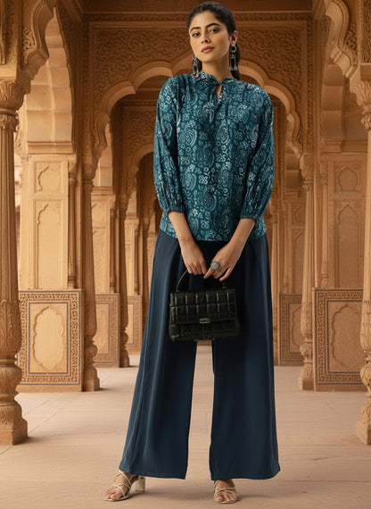 Woman in a teal patterned top and dark blue pants holding a black handbag in an ornate architectural setting. Blue Floral Printed Tie-Up Neck Top, Floral Printed Tunic, printed tunic, mandarin collar Tunic, casual wear Tunic, women’s tunic, Office Wear Tunic
